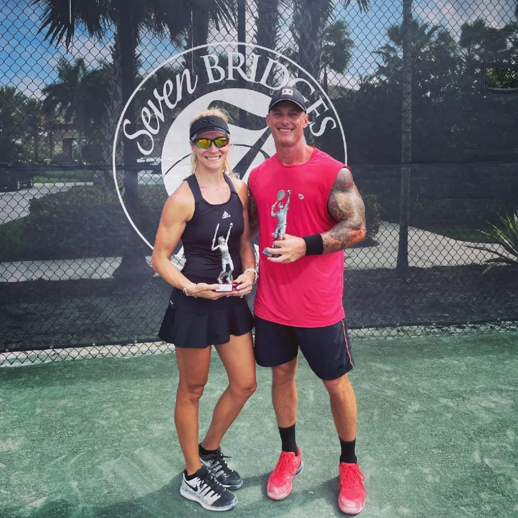 Two players holding tournament trophies in front of Seven Bridges Tennis branding